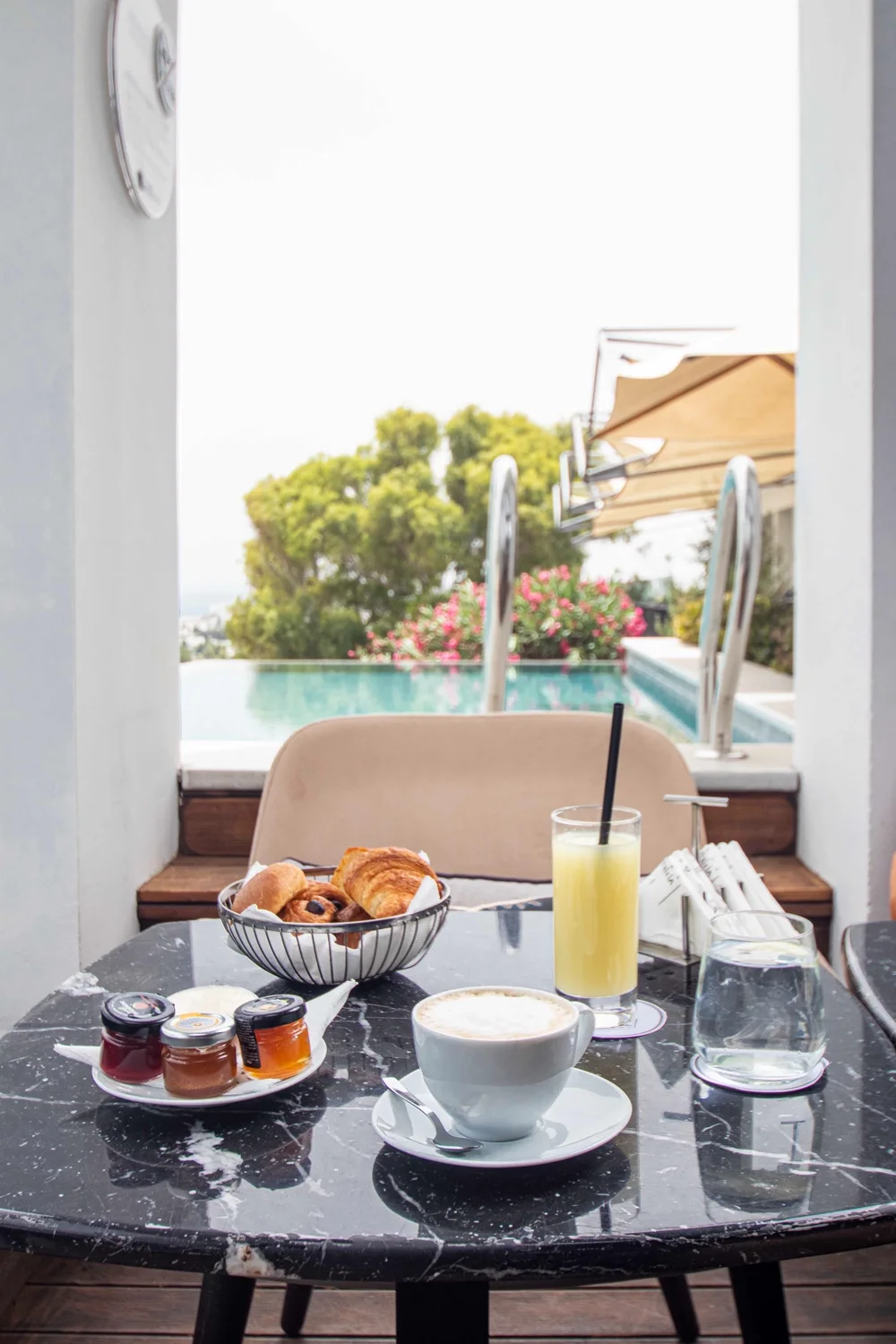 A basket of pastries, coffee and orange juice on a marble table, the pool behind