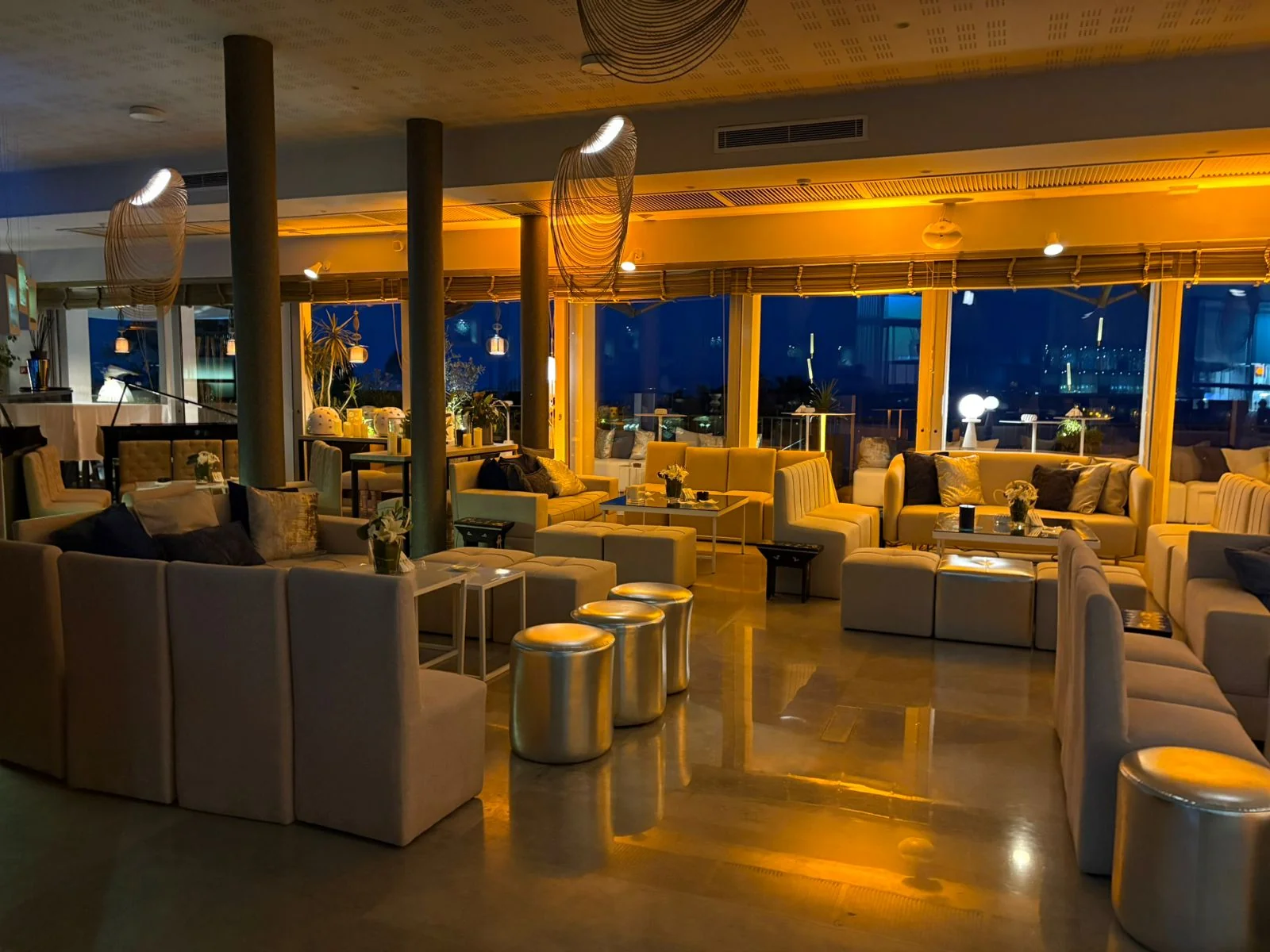 The dining room of Le RestÔ on a reception evening: pale sofas, silver poufs, golden light, the windows open to the night