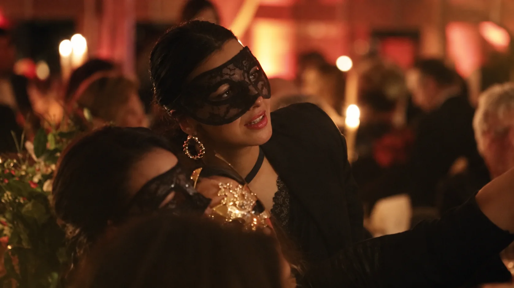 Two guests in black lace masks at a candlelit evening, in warm red and gold light