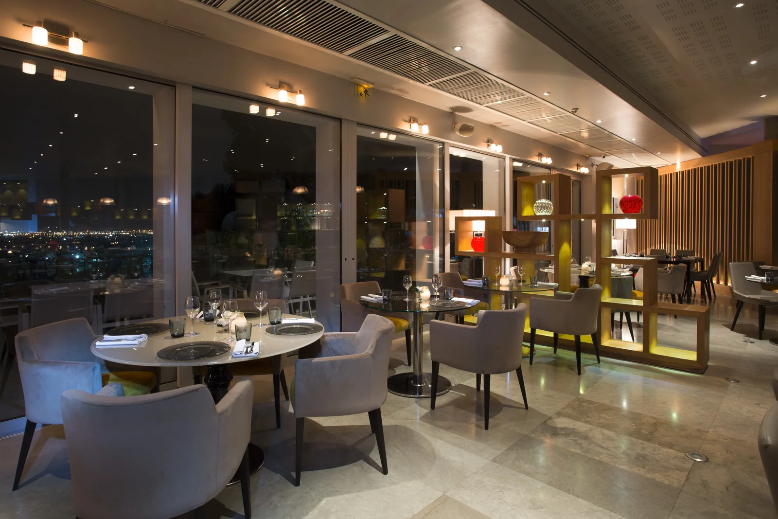 La salle du RestÔ le soir : tables dressées, fauteuils gris, étagères de bois éclairées et baies vitrées ouvertes sur les lumières de la ville