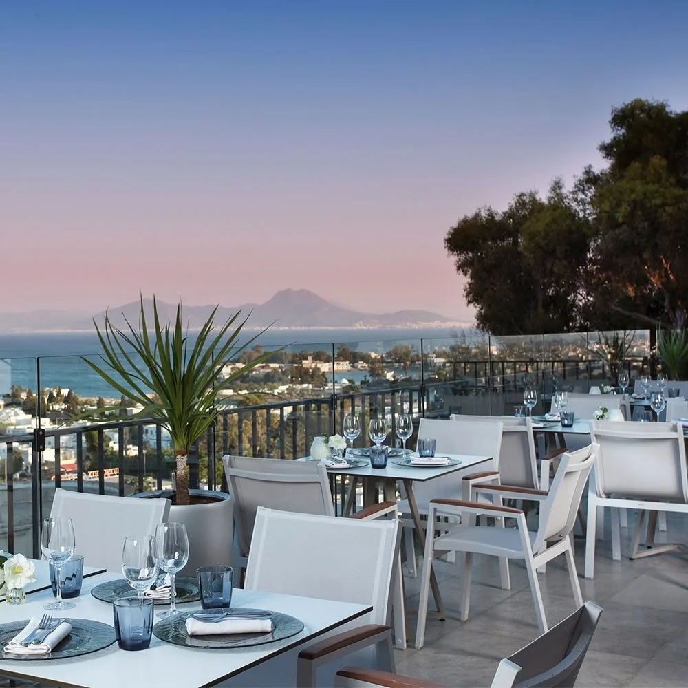 La terrasse au coucher du soleil : tables dressées, ciel rose, la baie de Carthage et le Boukornine au loin