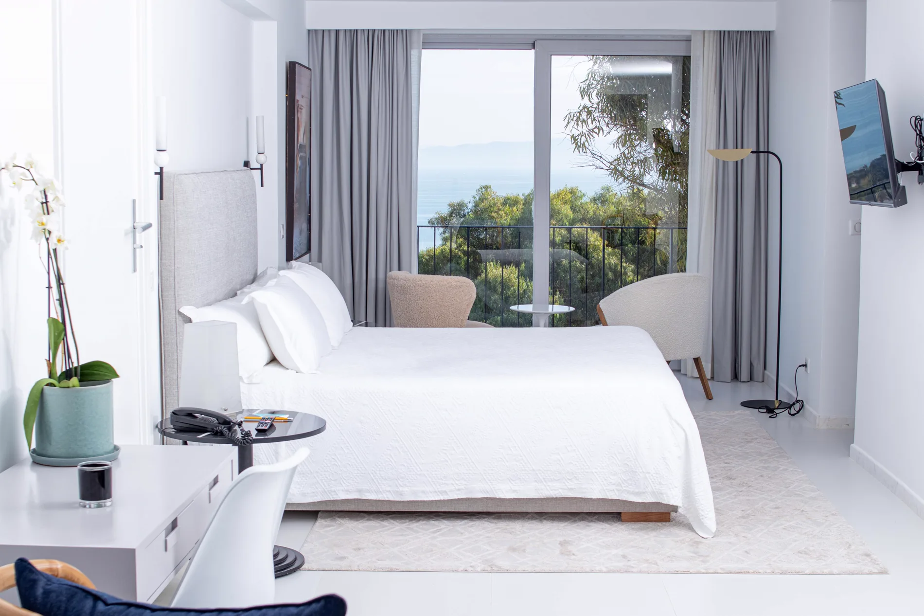 A suite at Villa Didon: white bed facing a picture window onto the sea and greenery, bouclé armchairs, soft daylight